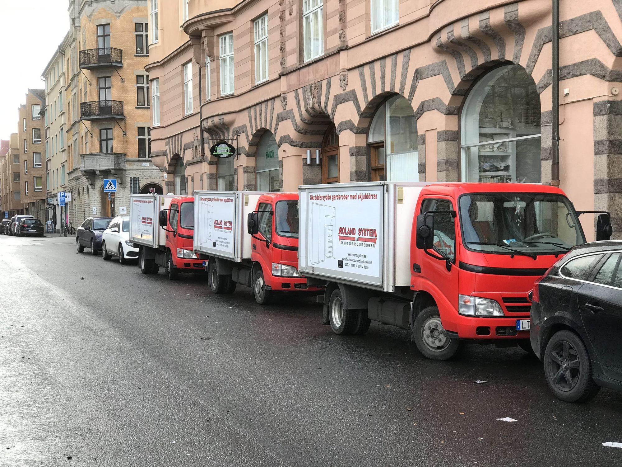 Three red Roland System trucks parked in a row on a city street in front of an older apartment building.