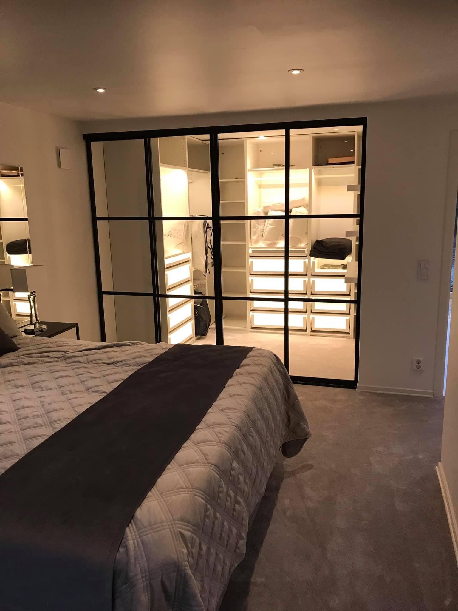 Black-framed glass wall with a lit walk-in wardrobe behind it, seen from a bedroom.