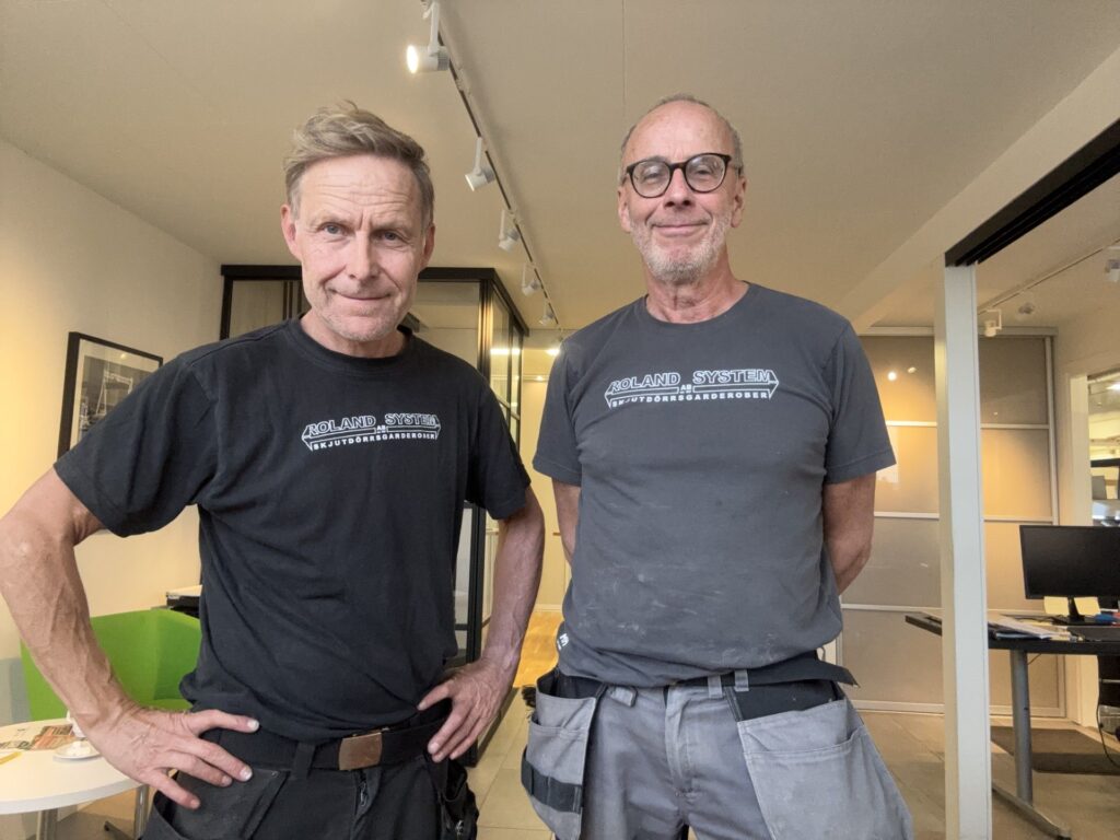 Two Roland System team members in work T-shirts with the company logo, smiling at the camera in front of black-framed glass walls.