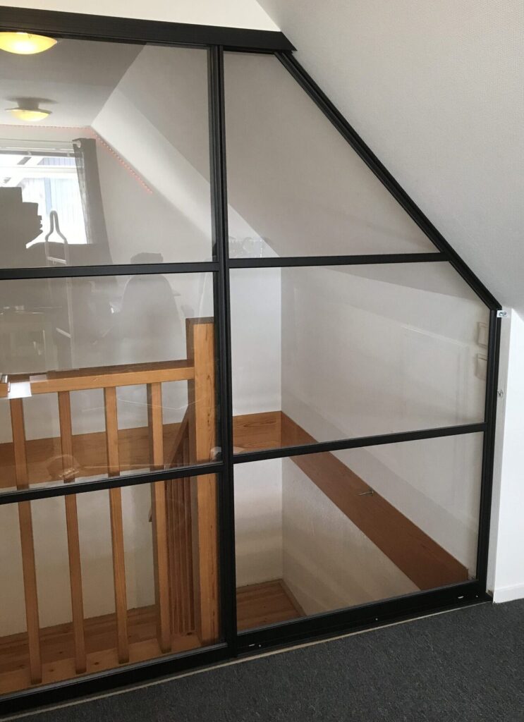 Black-framed glass wall under a sloped ceiling beside a stairwell with a wooden banister.